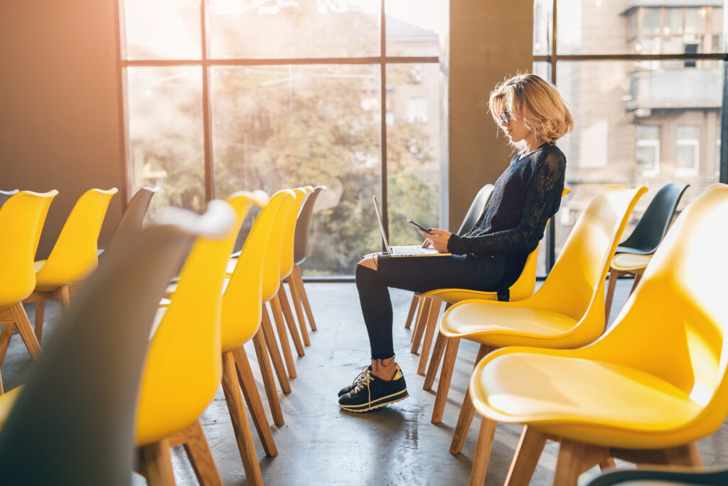 young pretty busy woman sitting alone in conference room, many yellow chairs, working at laptop, student education online, freelancer startup