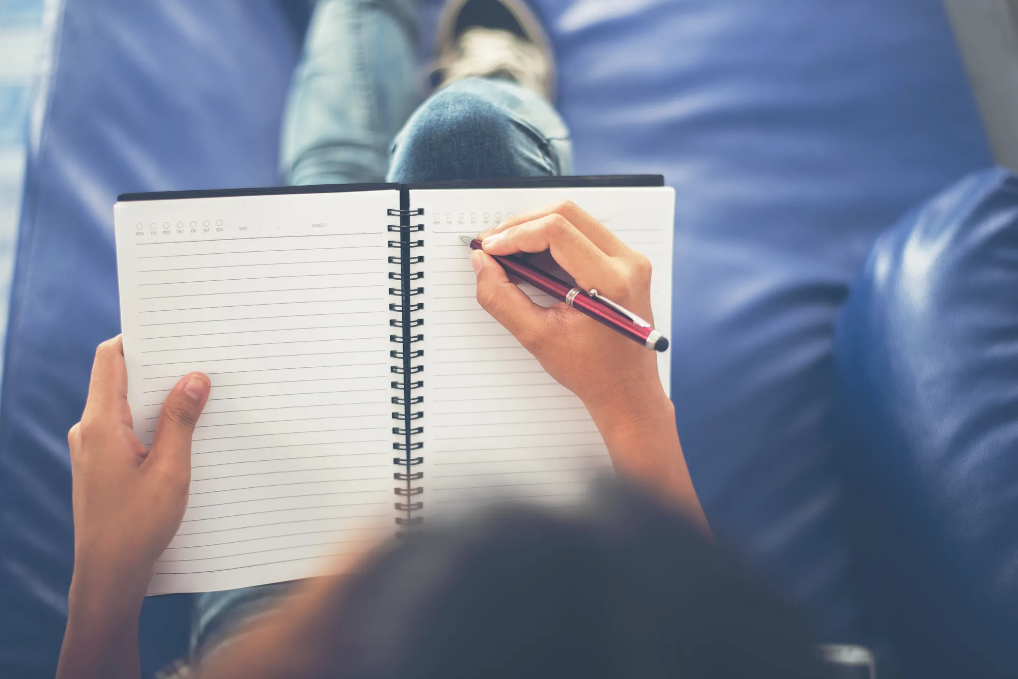 Overhead view of person writing in a spiral notebook on a blue cushion.