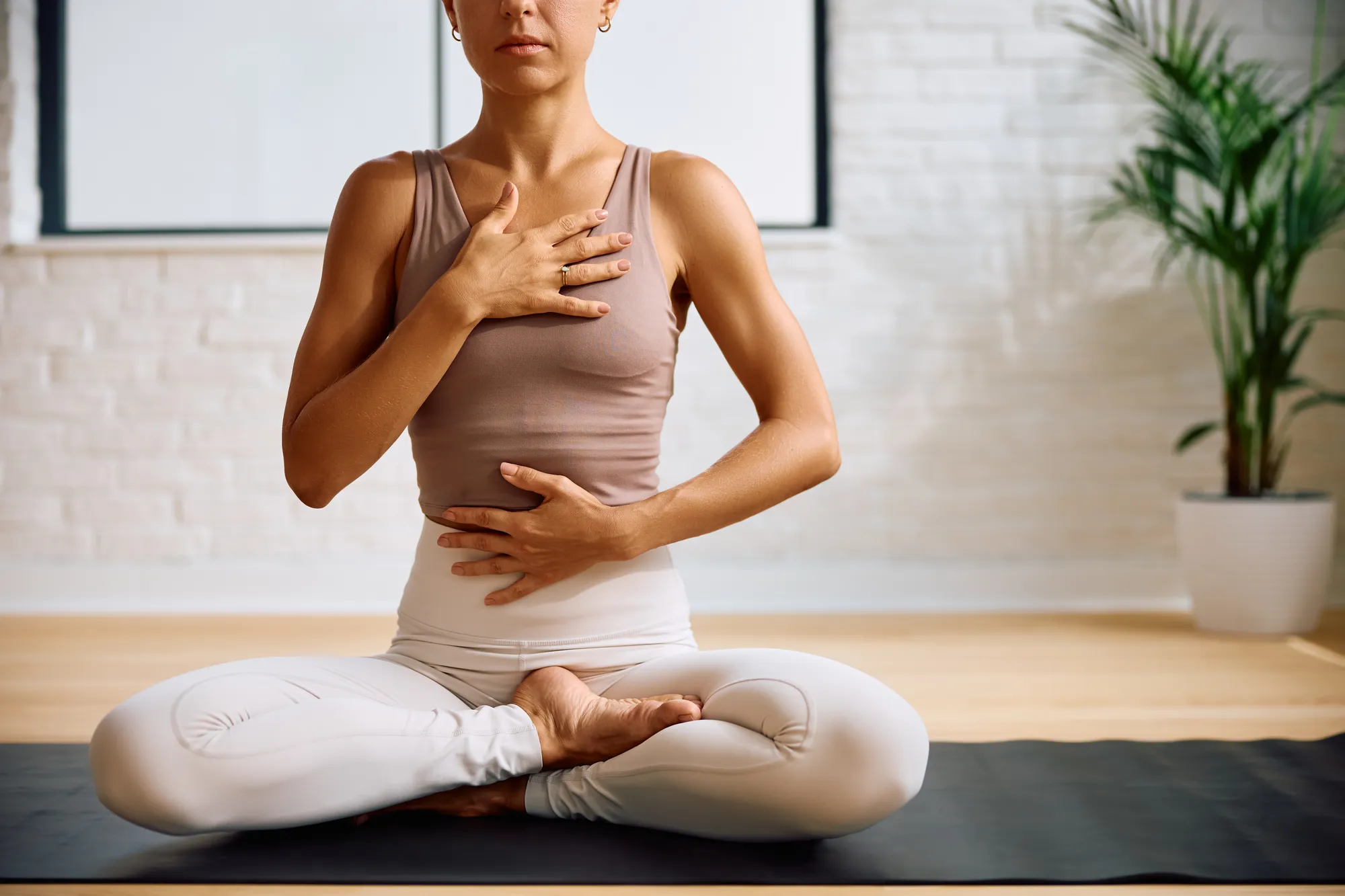Woman in lotus pose, hands on chest and abdomen, eyes closed, bright room.