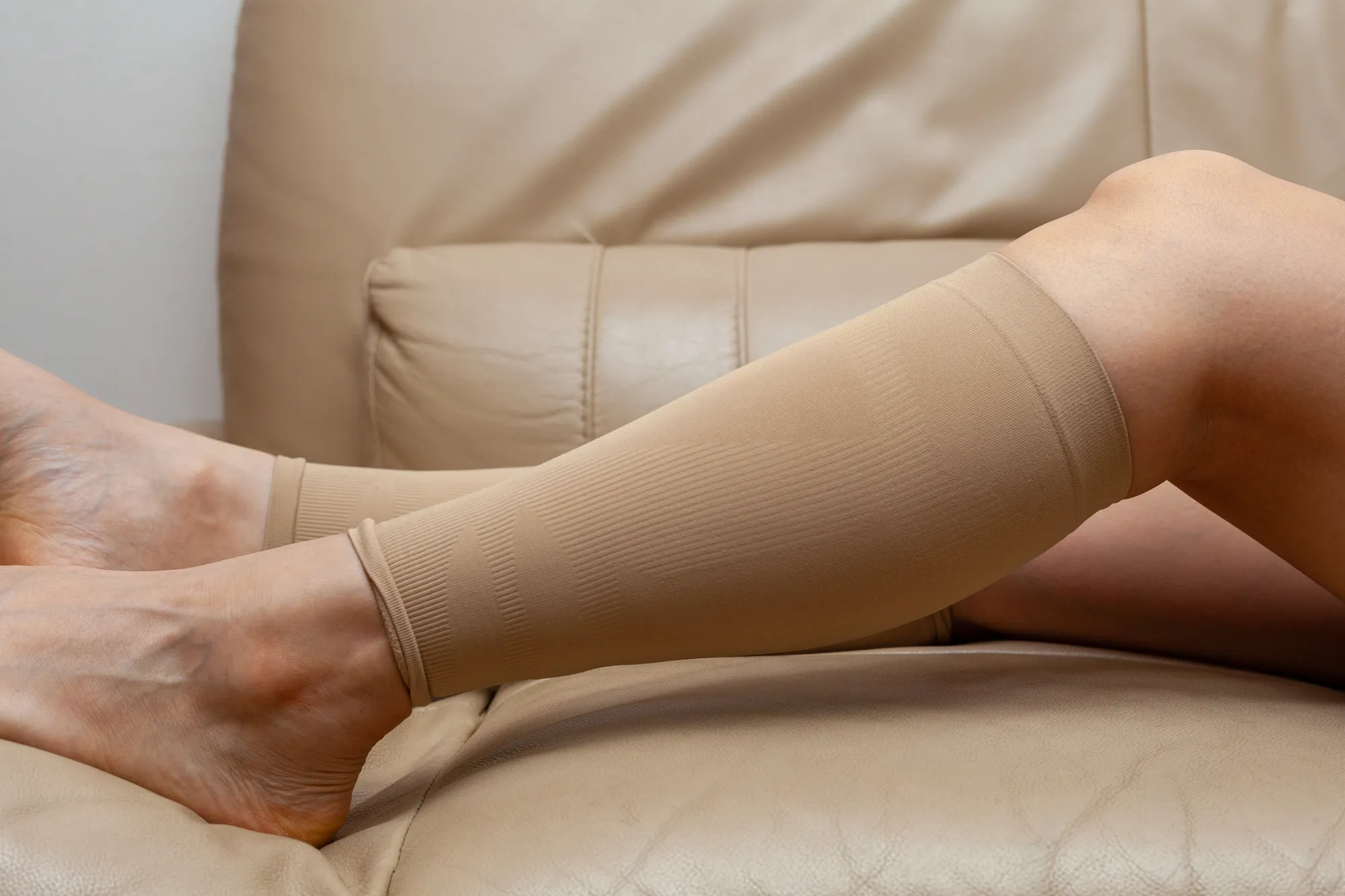 Legs in beige compression stockings resting on a tan leather sofa.