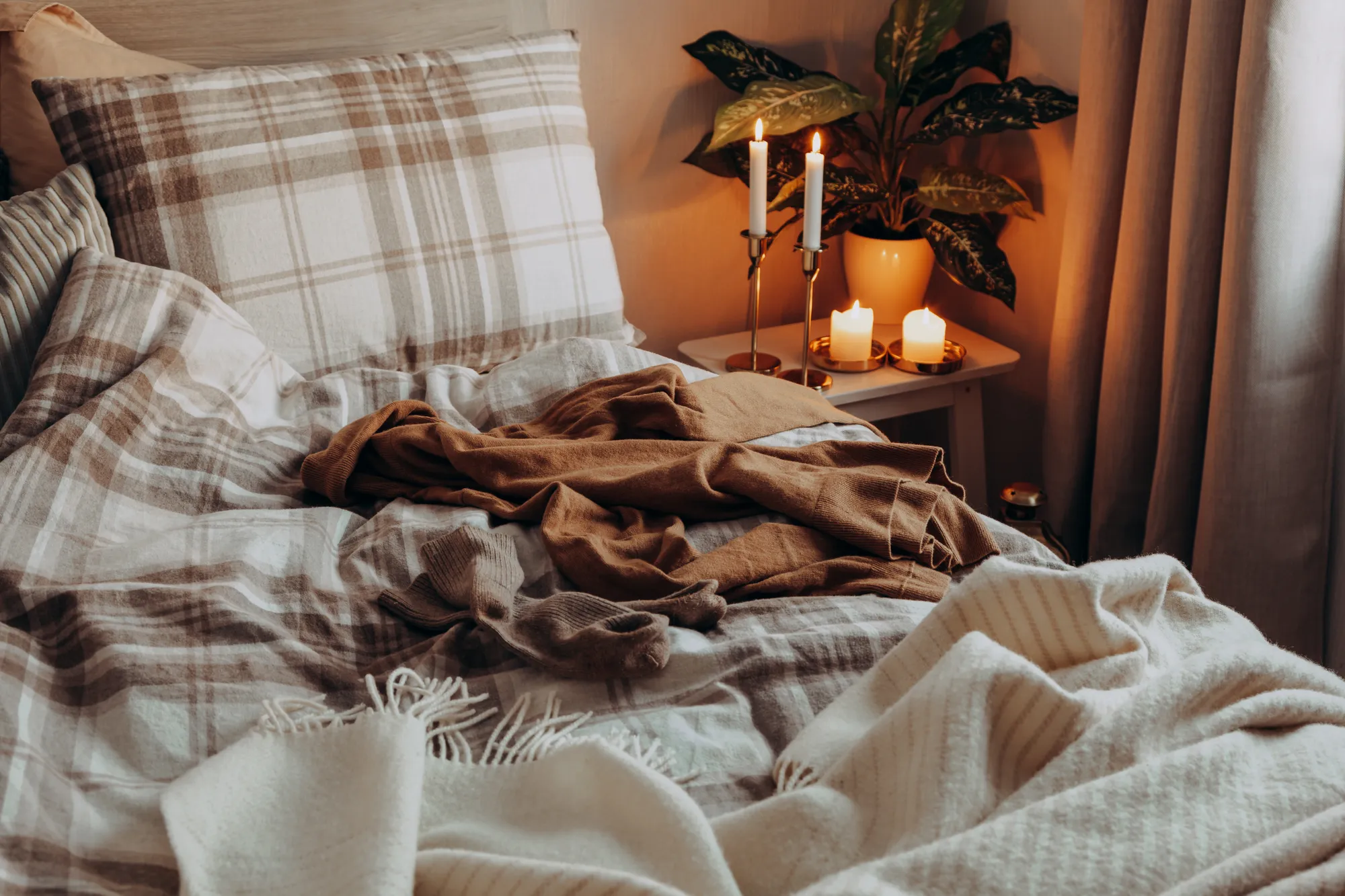 Cozy, unmade bed with a knit blanket and warm candles on a wooden bedside table.