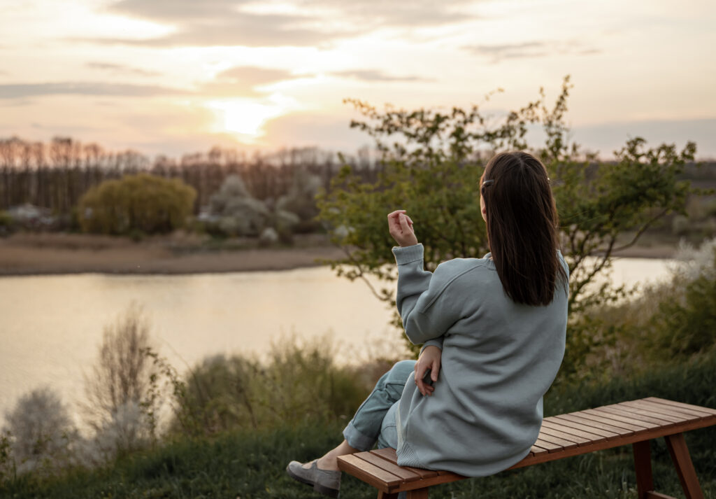 A girl in casual style sits and looks at the sunset near the lake.